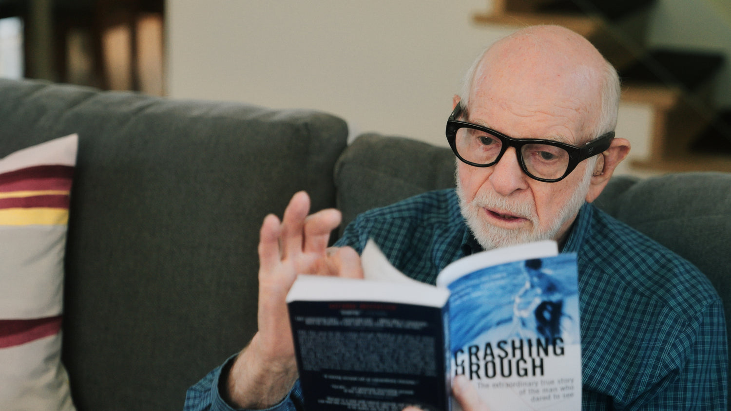 An older male user wearing Echo Vision smart glasses sits relaxed on a couch. One hand holds open the book 'Crashing Through' while the other hand flips a page, illustrating the device’s real-time reading capability.