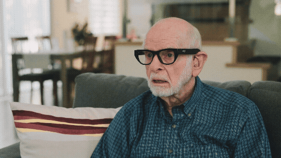 A video loop of an older male user on a couch wearing Echo Vision glasses, who smiles and waves. The perspective rapidly shifts to the glasses’ camera view, which interprets the scene by identifying a kitchen counter and a person standing behind it, illustrating the 'Remote Assistance' feature.
